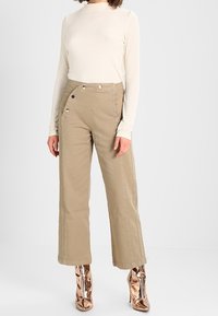 Mujer vistiendo pantalones beige de cintura alta con detalles de botones, una blusa ajustada de manga larga color crema, y botas de tobillo metálicas en color rosa dorado brillante.