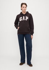 Hoodie GAP de culoare maronie cu litere albe, având un buzunar tip cangur. Asortat cu blugi negri și pantofi maroon. Design simplu și croială relaxată.
