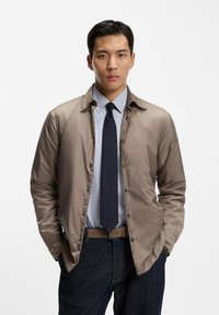 Giovane uomo che indossa una giacca color taupe abbottonata sopra una camicia azzurra con cravatta blu navy e jeans scuri, in piedi con le mani nelle tasche davanti a uno sfondo bianco.