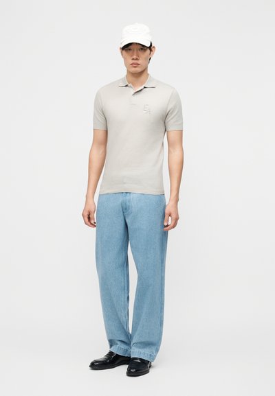 Άνδρας που φοράει ανοιχτό γκρι πόλο με κοντά μανίκια, ανοιχτό μπλε τζιν με φαρδιά μπατζάκια, μαύρα παπούτσια και λευκό καπέλο, στέκεται μπροστά σε απλό λευκό φόντο.