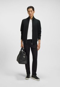 Svart zip-up jacka, vit skjorta, svarta byxor och svarta sneakers. Han håller en texturerad svart väska med en silverfärgad detalj.
