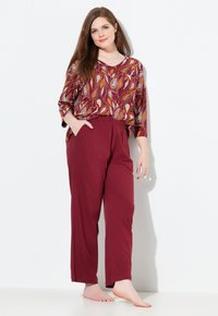 Langsleeve top met een paisleypatroon in burgundy en geel, gecombineerd met burgundy elastische broek. Model staat blootvoets tegen een witte achtergrond.