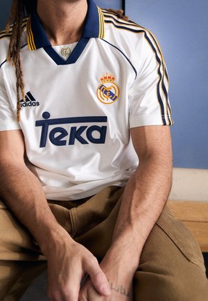 Persona con dreadlocks che indossa la maglia bianca del Real Madrid con dettagli blu navy e oro, seduta con le mani giunte in grembo.