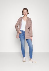 Double-breasted blazer in a brown and beige check pattern, paired with a white v-neck t-shirt and blue skinny jeans. Light sneakers.