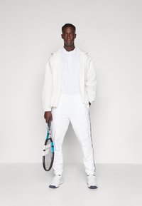 Vit texturerad jacka, vit t-shirt och vita träningsbyxor med svarta sidostripes. Håller en tennisracket och bär vita sportskor.