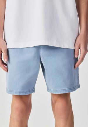 Person trägt hellblaue Shorts und ein weißes Shirt, steht entspannt mit an den Seiten hängenden Händen vor einem einfarbigen Hintergrund.