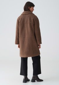 Brauner, strukturierter Fleece-Mantel mit Kragen, langen Ärmeln und mittellangem Saum. Getragen über schwarzen Hosen und Stiefeletten. Rückansicht.