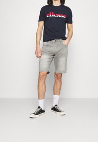 Man som bär en marinblå "ellesse" t-shirt, ljusgrå denimshorts, vita strumpor och svarta canvas sneakers, står med en hand i fickan.