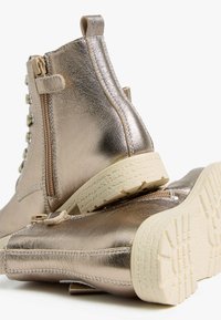 Bottines métalliques dorées avec une texture lisse, un laçage à l'avant, une fermeture éclair sur le côté et une semelle crème avec un motif de crampons.