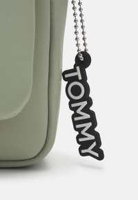 Borsa a mano verde in ecopelle con texture liscia, dotata di un'etichetta in gomma nera su cui è scritto "TOMMY" in lettere bianche, attaccata da una catena argentata.