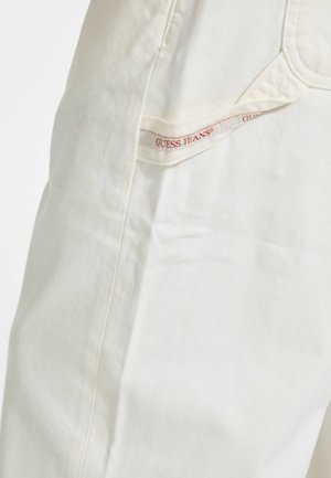 Jeans din denim alb cu o textură netedă, având o buzunar lateral vizibil cu un etichetă roșie "GUESS JEANS". Design simplu, detalii minime.