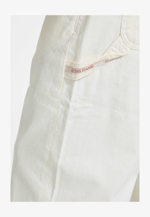 Bílé džíny ze silné denimové látky s hladkou texturou, se viditelnou boční kapsou a červeným štítkem "GUESS JEANS". Jednoduchý design, minimální detaily.