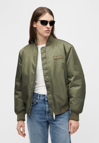 Άτομο με μακριά καστανά μαλλιά που φοράει μαύρα γυαλιά ηλίου, χακί μπουφάν τύπου bomber με το λογότυπο "Off-White", λευκό πουκάμισο και μπλε τζιν.