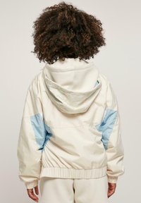 Witte en lichtblauwe windbreaker jas met capuchon, elastische manchetten en tailleband. Dichte textuur, oversized pasvorm en contrasterende kleurelementen.