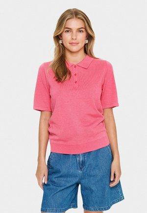 Polo shirt rose à manches courtes avec un col, présentant une patte de boutonnage à trois boutons et un ourlet côtelé. Associé à un short en denim bleu.
