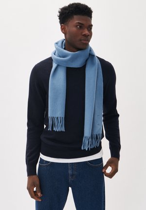 Écharpe bleu clair en tissu doux, avec une finition texturée et des bouts frangés. Portée sur un pull sombre devant un arrière-plan uni.