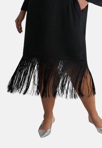 Černé šaty s třásňovým lemem, které zdobí dlouhé černé třásně. Modelka má na nohou stříbrné baleríny. Šaty jsou hladké, s volným střihem a kapsami.