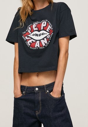 Vrouw draagt een zwart cropped T-shirt met een rood-wit Pepe Jeans London-logo, gecombineerd met donkerblauwe spijkerbroek, handen in de zakken.