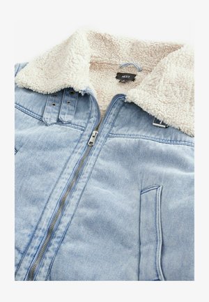 Chaqueta de denim con cuello forrado de sherpa, de color azul claro, con cremallera frontal y detalles de bolsillo. Etiqueta visible en la zona interior del cuello.