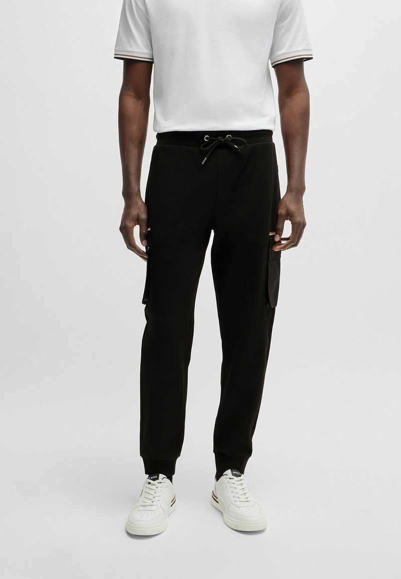 Pantalon de jogging noir avec taille élastique et cordon de serrage, comportant des poches latérales et des poignets côtelés. Associé à un polo blanc.
