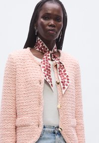 Rózsaszín, texturált tweed dzseki arany gombokkal, két zsebbel. Fehér felsővel és nyaknál megkötött virágmintás sállal párosítva.