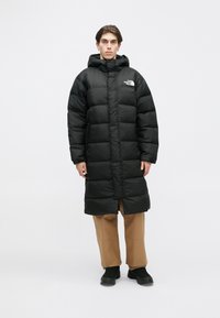 Crna jakna na preklop s kapuljačom, prošiveni dizajn, prednji patentni zatvarač i logo na lijevoj strani prsiju. Nosit će se preko smeđih hlača i crne obuće.