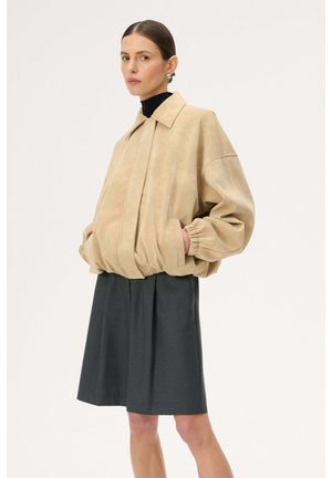 Kobieta w beżowej, oversize'owej kurtce z elastycznymi mankietami, na czarnym golfie i grafitowej plisowanej spódnicy, z rękami w kieszeniach, na neutralnym tle.