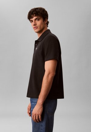 Joven con camiseta tipo polo negra de manga corta y vaqueros azules de pie de lado frente a un fondo gris liso.