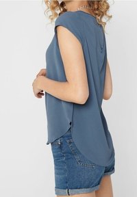 Haut bleu à manches courtes avec un col rond, présentant un ourlet arrondi et une petite fente dans le dos, associé à un short en denim. Tissu texturé.