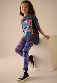 T-shirt graphique gris avec un design coloré de Stitch, associé à des leggings floraux vibrants en bleu, rose et violet, et des baskets montantes noires.