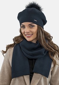 Beret tricotat negru, cu un pompon pufoș pe partea de sus, asortat cu o eșarfă închisă la culoare. Ambele au un finisaj texturat și margini canelate.