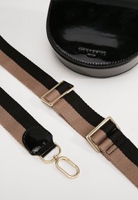 Kulatá taška z černé lakované kůže s lesklým povrchem, doplněná dvojitým pruhovaným popruhem v černé a světle hnědé barvě, se metalovými kovovými prvky.