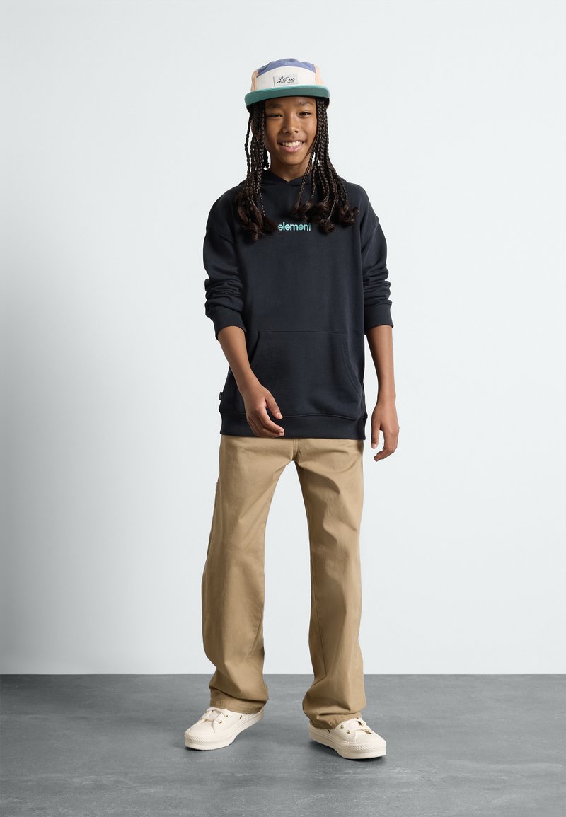 Dunkelblaue Kapuzenjacke mit einer Fronttasche, beiger lässiger Hose und weißen Turnschuhen. Eine mehrfarbige Mütze mit einem Logo rundet das Outfit ab.