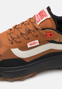Rudos Vans sportbačiai su tekstūruotu audiniu, juodais akcentais, balta šonine juostele ir storu guminiu padu. Raudonas logotipo etiketas ant liežuvėlio.