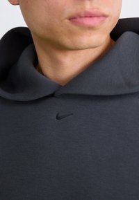 Nike hoodie lavet af mørkegråt stof, med en stor hætte, en struktureret overflade, og et sort broderet logo på brystet.