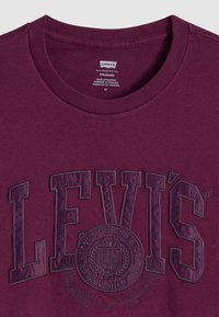 Burgunderfarbenes T-Shirt mit kurzen Ärmeln und strukturiertem, dunklem Schriftzug, das "Levi's" und ein kreisförmiges Logodesign in der Mitte zeigt.