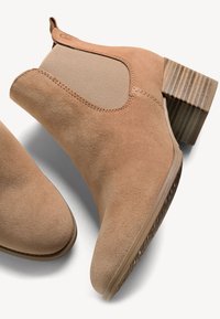 Botas de tobillo de ante en un color marrón claro, con paneles laterales elásticos, punta redondeada y un tacón de madera apilada con superficie texturizada.