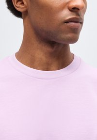 Κοντινό πλάνο του κάτω μέρους του προσώπου και του λαιμού ενός ατόμου που φοράει ένα ανοιχτό μοβ μπλουζάκι με στρογγυλή λαιμόκοψη σε απλό φόντο.