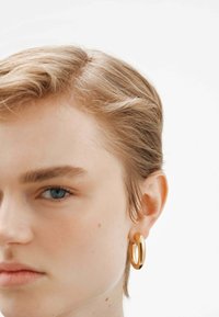 Los pendientes de aro en tono dorado presentan un diseño suave y redondeado con una superficie pulida, exhibidos en la oreja de un modelo sobre un fondo liso.