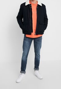 Mörkblå bomullsjacka med vit fleecekrage, parat med en orange sweatshirt och blå jeans; modellen bär vita sneakers.