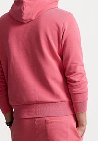 Felpa rosa realizzata in tessuto morbido, con cappuccio, maniche lunghe, polsini e orlo a costine, e un piccolo dettaglio di tasca posteriore.