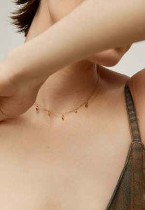 Collar de oro con cinco pequeños colgantes en forma de péndulo. La cadena es delicada, con una textura sutil que complementa un hombro desnudo y tirantes de denim.