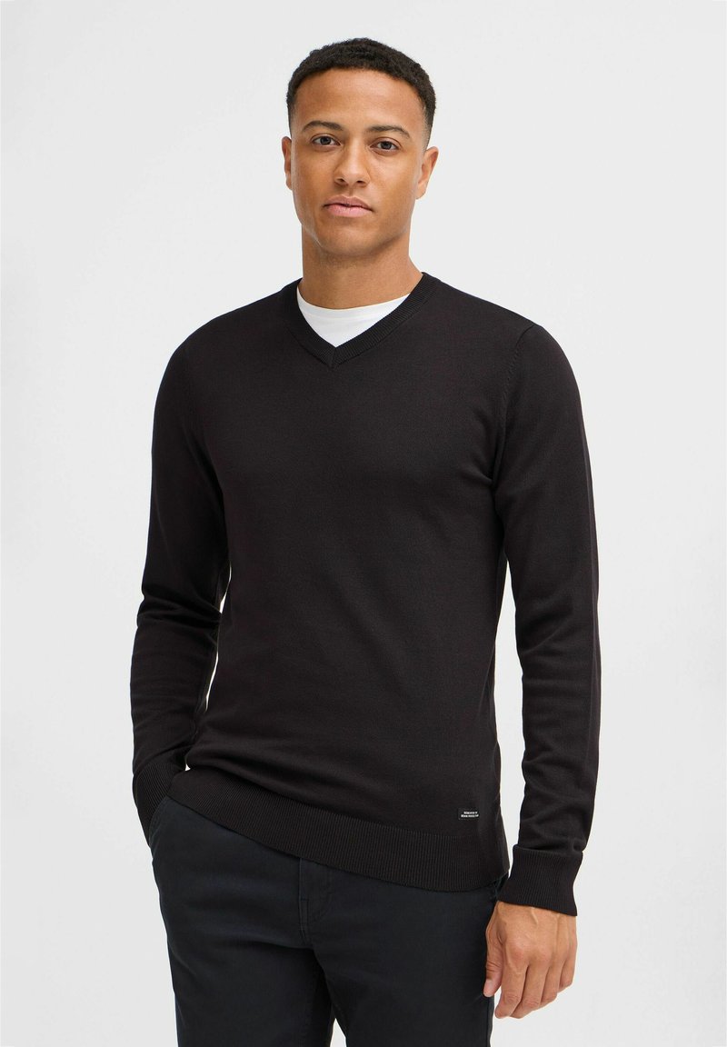 Pulover negru cu decolteu în V, cu mâneci lungi, design potrivit, cu manșete și tiv ribate, realizat dintr-un material tricotat neted, purtat peste un tricou alb.