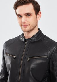 Zwarte leren motorjack met een hoge kraag, gecapitonneerde schouderaccenten en twee voorzakken met rits en zilverkleurige hardware.