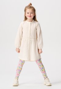 Jeune fille aux cheveux blonds en chignon, portant une robe crème à œillets, des leggings fleuris et des baskets blanches, se tenant avec les jambes écartées sur un fond blanc.