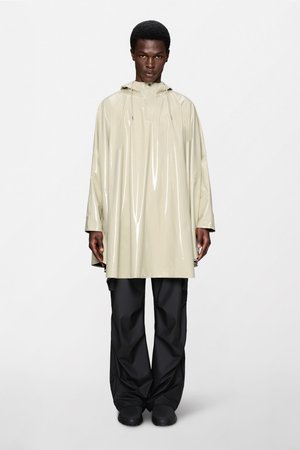Mužský model oblečený v lesklom béžovom pršiplášti s kapucňou a predným zipsom, voľné čierne nohavice a čierne topánky, stojaci pred jednoduchým pozadím.