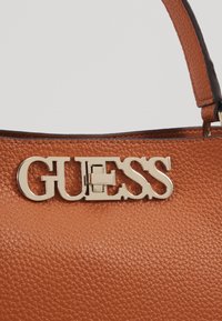 Brązowa torba o teksturowanej powierzchni z wyróżniającym się złotym logo "GUESS" jako zapięcie, z ciemnym paskiem dla dodatkowego akcentu.