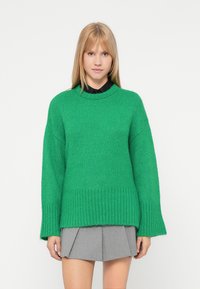 Πράσινο πλεκτό πουλόβερ με στρογγυλή λαιμόκοψη, ριμπ μανίκια και στρίφωμα. Απλός, μεγάλος σχεδιασμός σε συνδυασμό με μια γκρίζα πλισέ φούστα.