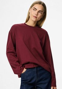 Młoda kobieta z prostymi blond włosami nosi luźną, bordową koszulę z długim rękawem oraz ciemnoniebieskie dżinsy, pozując z jedną ręką w kieszeni.