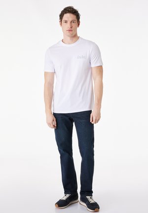 Mężczyzna stoi, ubrany w białą koszulkę z krótkim rękawem z małym napisem na klatce piersiowej, ciemnoniebieskie dżinsy i czarne trampki z białymi sznurówkami.